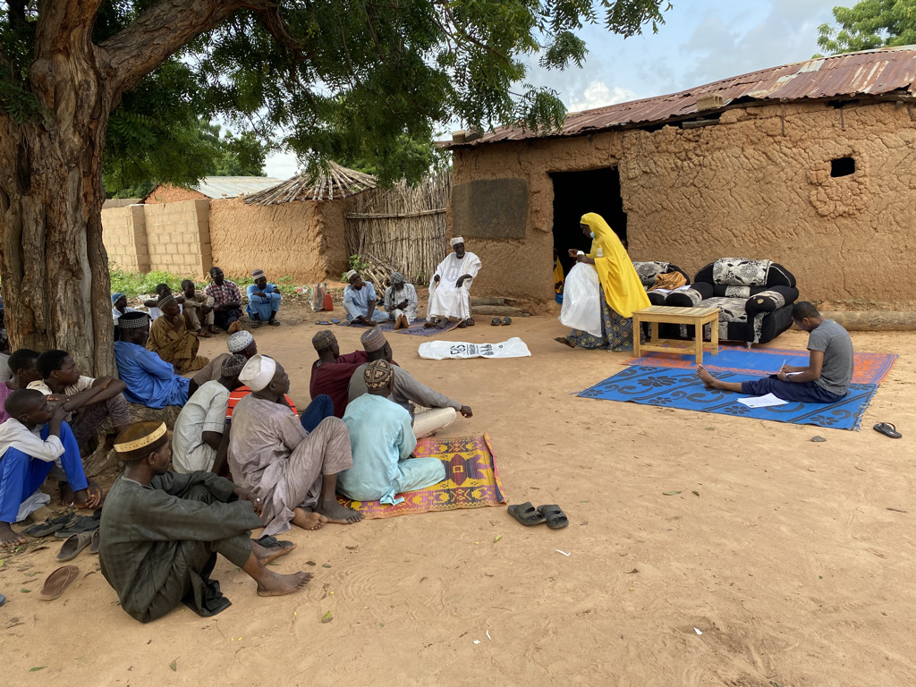 group of people sitting on the floor under a tree listening to a person