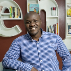 Aaron Kirunda smiling at camera wearing blue and white checked button down shirt, in front of bookcases shaped like letters E and N