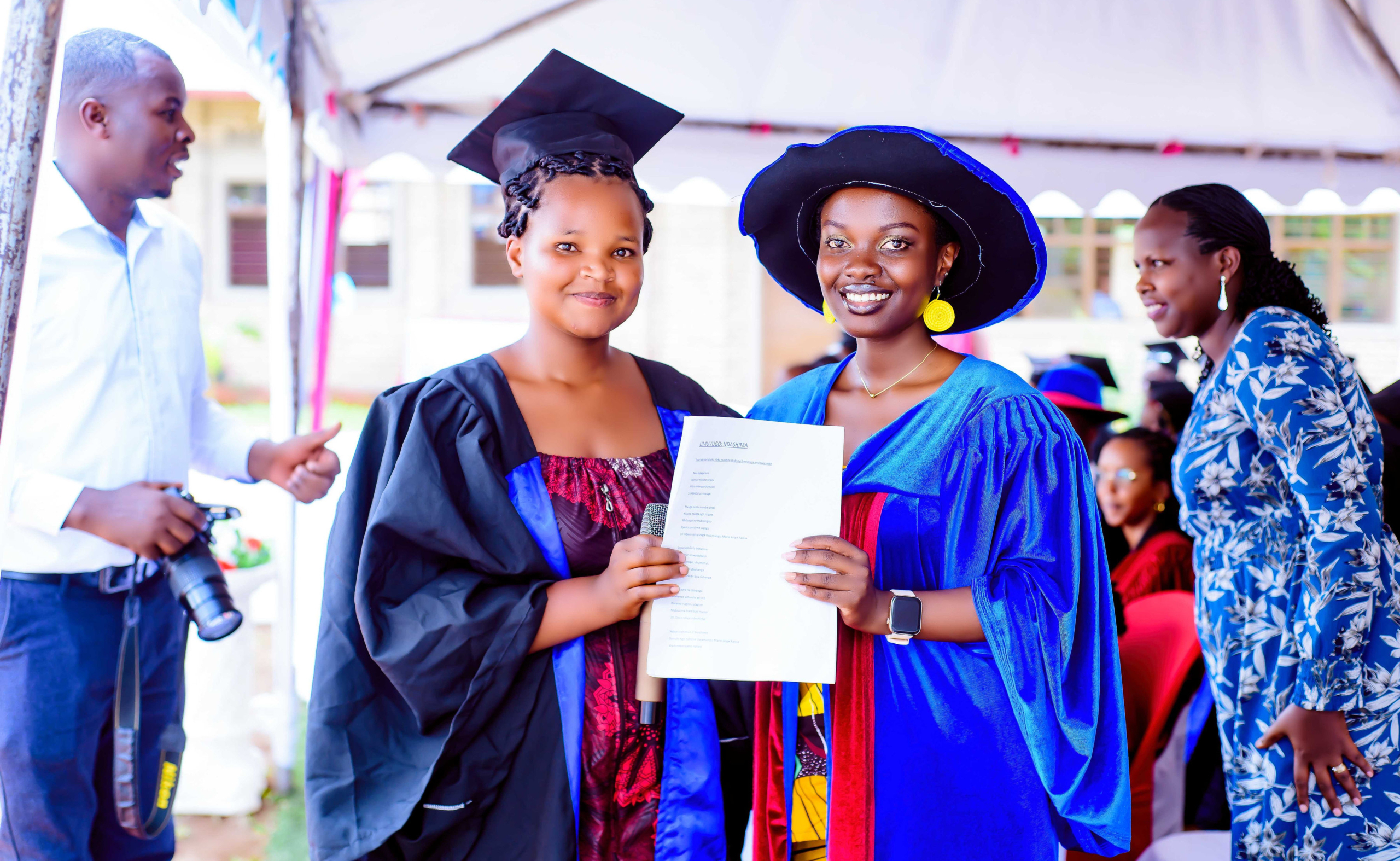 Female graduate of Impanuro wearing graduation gown and cap smiling getting handed diploma by another woman in gown and hat