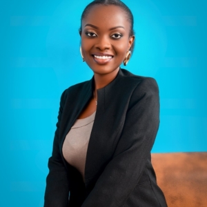 Raissa Uwamungu smiling at camera, seated in front of blue backdrop wearing black blazer over grey shirt, with hair pulled back neatly