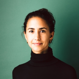 Rhoda Berger wearing black turtleneck with hair tied back in bun in front of green backdrop