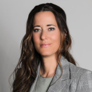 Profile Picture Marion Gehring softly smiling at camera with brown hair falling over right shoulder, wearing grey blazer in front of grey backdrop